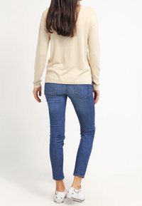 Beige Langarmoberteil kombiniert mit blauen Skinny-Jeans. Die Jeans haben eine verwaschene Optik und Rückentaschen. Weiße Sneakers runden das Outfit ab.