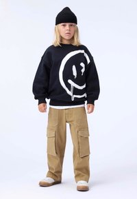 Svart genser med stort hvitt smilefjesgrafikk, parret med brune cargo-bukser med flere lommer, og hvite sneakers. Svart lue.