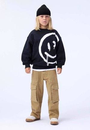 Svart genser med stort hvitt smilefjesgrafikk, parret med brune cargo-bukser med flere lommer, og hvite sneakers. Svart lue.