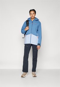 Chaqueta impermeable azul y celeste con capucha, frente con cremallera y dos bolsillos laterales. Combinada con pantalones oscuros y zapatos beiges.