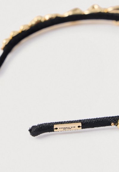Czarna opaska na głowę z ozdobnym złotym obszyciem i małą złotą etykietą z wygrawerowanym napisem "EUGENIA KIM NEW YORK" na jednym końcu.