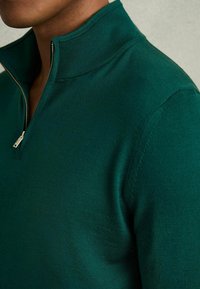 Pull vert ajusté avec un col zippé, fabriqué en maille lisse. Il présente un col côtelé et une texture subtile sur le tissu.