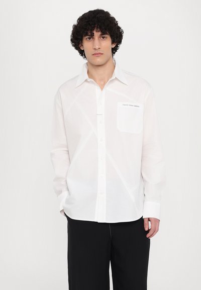 Camisa blanca de botones confeccionada en una tela ligera, con un bolsillo frontal, detalles de costura diagonal y un cuello clásico. Pantalones negros.