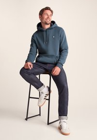 Hooded sweatshirt in teal cotton blend, featuring a front pocket and logo on the chest. Paired with dark denim jeans and white sneakers.