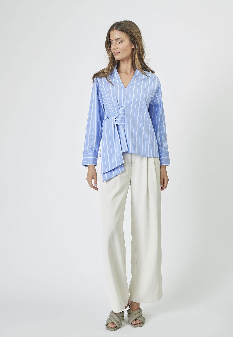 Light blue wrap blouse with vertical white stripes, long sleeves, and a front tie detail, paired with white wide-leg trousers.