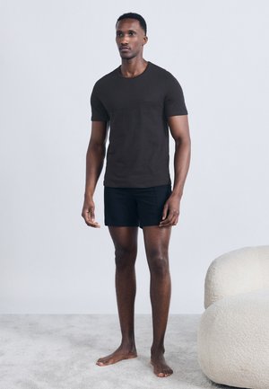 Uomo in piedi a piedi nudi su un tappeto, indossa una maglietta nera a maniche corte e pantaloncini neri, con una poltrona bianca curva nelle vicinanze.