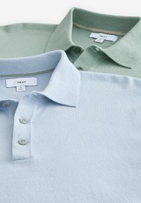 Zwei poloshirts mit Kragen übereinander gestapelt: eines in Hellblau, das andere in sanftem Grün. Beide verfügen über einen strukturierten Strickstoff und Knopfverschlüsse. Markenetikett sichtbar.