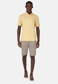 Polo tričko v svetložltej bavlne s trojradovým gombíkovým zapínaním, skombinované so sivými šortkami. Hnedé slip-on topánky dotvárajú outfit.