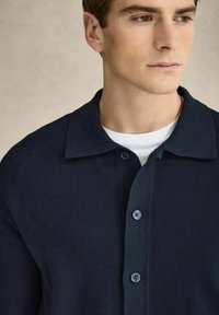 Navy ribbed knit cardigan with a spread collar and buttoned front, featuring subtle texture and round buttons. White undershirt visible.