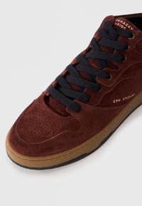 Brown suede high-top sneaker with black laces, perforated toe box, and gum sole. Features "CPH STUDIO" in gold on the side.