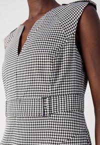 Robe sans manches pied-de-poule noir et blanc avec col en V et large ceinture assortie avec une boucle rectangulaire portée par une personne à la peau foncée.
