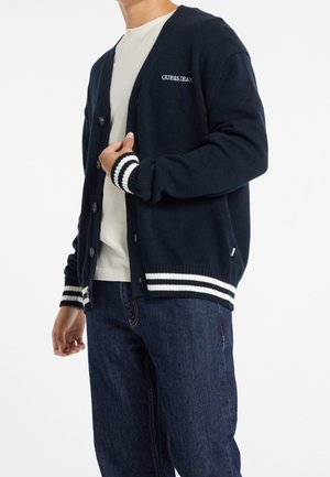 Hombre con cárdigan azul marino con rayas blancas y logotipo "Guess Jeans", camiseta blanca y vaqueros azul oscuro sobre fondo blanco.