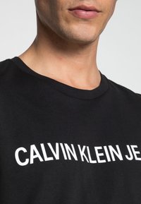 Detailní záběr na osobu v černém tričku s bílým textem, který částečně zní "CALVIN KLEIN JE" na hrudi.
