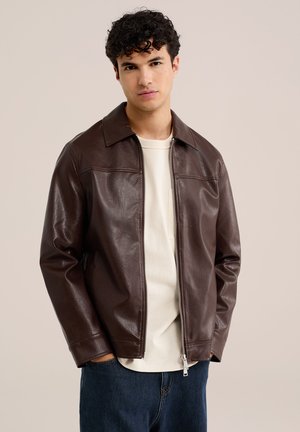 Mann mit lockigem Haar, der eine dunkelbraune Lederjacke über einem weißen Hemd und blauen Jeans trägt, steht mit den Händen in den Taschen vor einem einfarbigen Hintergrund.