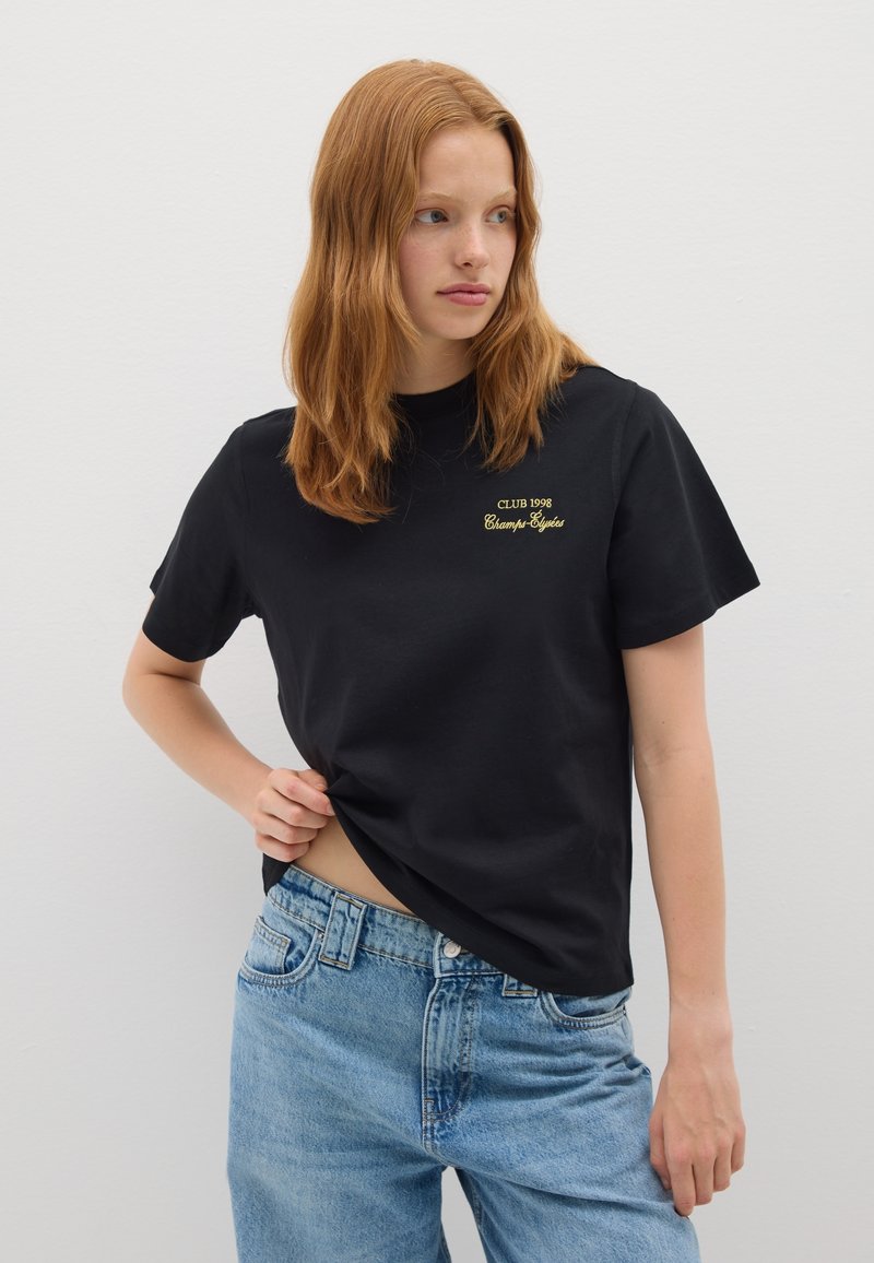 Camiseta de algodón negra con cuello redondo, mangas cortas y pequeño texto bordado en dorado: "CLUB 1998 Champs Styles" en el pecho.