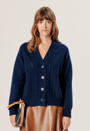 Femme aux cheveux bruns mi-longs portant un cardigan en maille bleu marine avec des boutons et une jupe en cuir marron, tenant un petit sac.