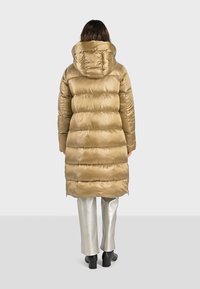 Doudoune longue et bouffante en tissu doré brillant avec une capuche, présentant des coutures horizontales et des fermetures éclair sur les côtés. Portée sur un pantalon argenté et des bottes noires.
