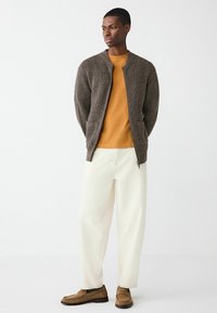 Brauner gerippter Cardigan mit zwei Fronttaschen, getragen über einem senfgelben Shirt, kombiniert mit übergroßen weißen Hosen und beigen Loafers.