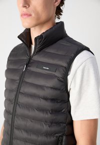 Schwarze, puffy Weste mit hohem Kragen, horizontalen Nähten und Reißverschluss. Mit einem kleinen Calvin Klein-Logo auf der Vorderseite. Getragen über einem grauen Hemd.