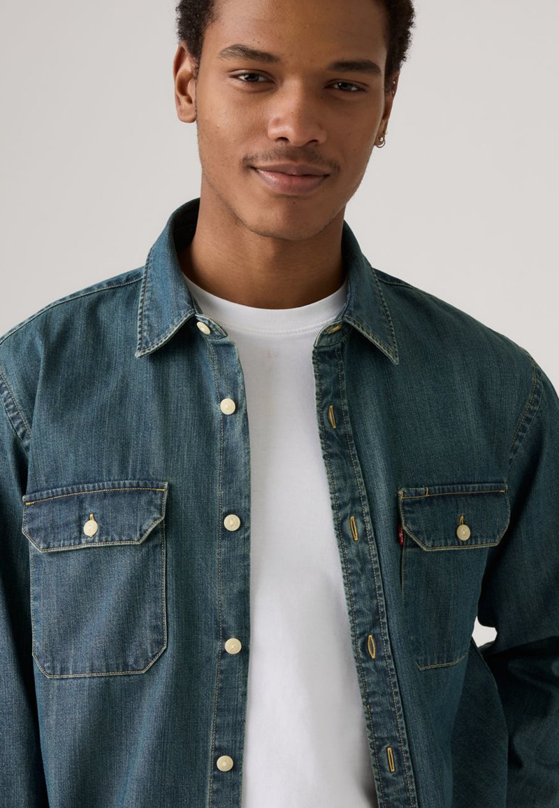 Camicia di jeans blu medio con chiusura a bottoni, due tasche sul petto e bottoni bianchi. Presenta un colletto classico e una texture morbida.