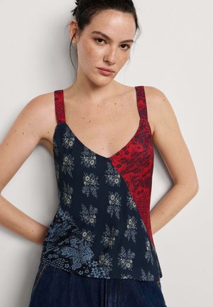 Frau mit dunklen Haaren, die ein ärmelloses Top mit roten floralen und blauen gemusterten Bereichen trägt, steht vor einem einfarbigen weißen Hintergrund.