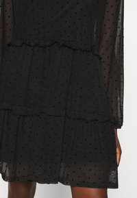 Schwarzes Kleid aus transparenter Stoff mit Polka-Dot-Muster, Rüschendetails, gestaffeltem Design und langen Ärmeln. Verstellbare Taille.