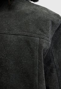 Svart mockafiber som visar en strukturerad, borstad yta. Har raka sömmar och en krage, med fibrer synliga genom hela materialet.