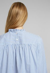 Personne blonde vue de dos portant une chemise à rayures verticales bleu et blanc avec col volanté et deux boutons blancs au centre.