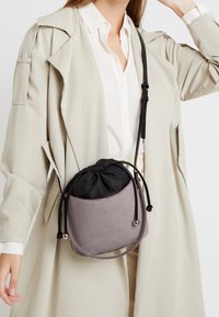 Szara teksturowana torba z marszczonym czarnym górnym wykończeniem, czarnym ściągaczem i srebrnymi akcentami. Noszona na skos przez jasnobeżowy płaszcz.