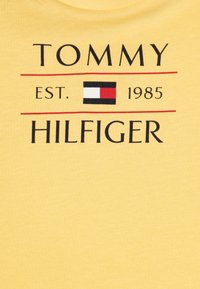 Żółta bawełniana koszulka z nadrukiem "TOMMY HILFIGER" w pogrubionym czarnym tekście, z kolorowym prostokątnym logo i rokiem założenia 1985.