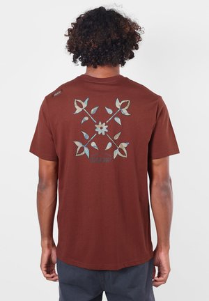 Personne aux cheveux bouclés portant un t-shirt marron avec un motif floral bleu et beige et le mot OXBOW dans le dos sur fond blanc.