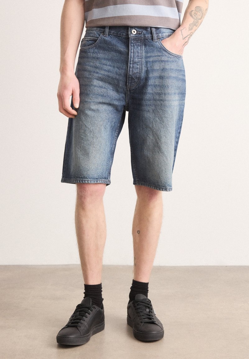 Shorts en jean bleu délavé, avec une coupe décontractée, un design à cinq poches et un léger effet de délavage. Associés à des baskets noires et des chaussettes.
