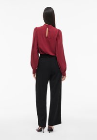 Bordeauxfarbige, strukturierte Bluse mit puffartigen Ärmeln und Schlitz im Rücken, kombiniert mit schwarzen, weiten Hosen und Absatzschuhen.