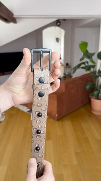 Cinto em pele castanha com um padrão de manchas circulares claras e escuras. Apresenta uma fivela de metal preta e tachas decorativas pretas.