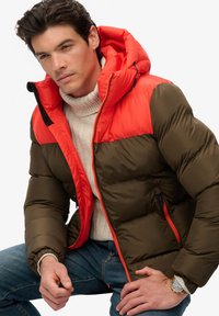 Puffjacke mit einem olivgrünen Körper und einem leuchtend roten oberen Abschnitt, einer Kapuze, strukturiertem Stoff, seitlichen Taschen und kontrastierenden Reißverschlüssen.