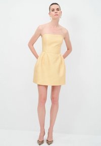 Robe jaune sans bretelles avec une finition texturée, corsage ajusté et jupe évasée, avec des poches latérales et portée avec des talons aiguilles dorés ornés.