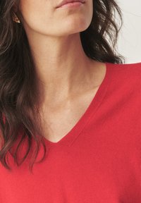 Žena s tmavými vlnitými vlasy, která má na sobě malé zlaté náušnice a červený svetr s V-výstřihem, viditelná od brady po horní část hrudníku.
