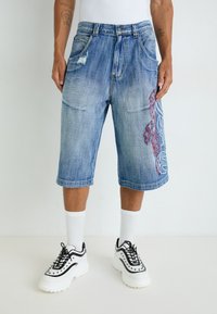 Jeansshorts mit einem hellblauen Waschung, versehen mit abgenutzten Stellen und rotem, filigranem Stickerei auf der linken Seite. Kombiniert mit klobigen schwarzen Turnschuhen.