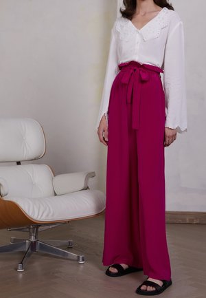 Blusa blanca con un escote de encaje, combinada con pantalones magenta de pierna ancha atados en la cintura y sandalias negras.