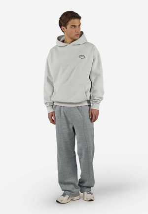 Jonge man met een lichtgrijze hoodie, grijze geplooide broek en witte sneakers, staand met handen langs zijn zij en naar rechts kijkend.