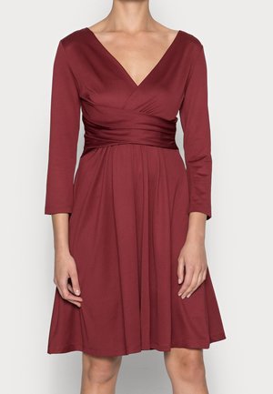 Kvinde iført en knælængde, langærmet burgunderfarvet kjole med V-hals og detaljer ved den rynkede talje.