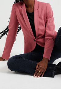 Růžový blazer s přesným střihem, zapínání na jeden knoflík a klopové límce. Černý top a tmavé džíny doplňují celý outfit.