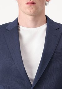 Tamno plavi sako s suptilnom karirastom teksturom, s reverima u obliku slova V i bijelom majicom s okruglim izrezom ispod. Usredotočen pogled na područje ovratnika.
