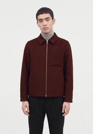 Ung mand iført en vinrød lynlåsjakke med brystlomme og en grå rullekravesweater, stående foran en ensfarvet hvid baggrund.