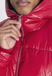 Rote, glänzende Puffjacke mit Reißverschlussfront und Kapuze. Verfügt über eine glatte Textur und gerippte Bündchen. Hand greift den Reißverschluss.