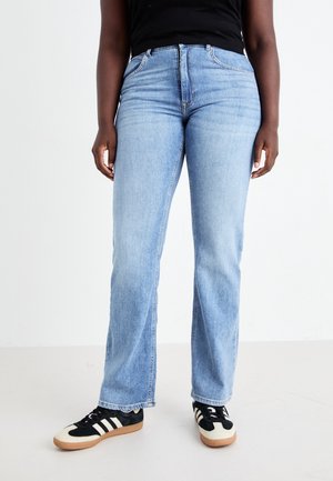 Personne portant un jean droit bleu clair, un haut noir et des baskets noir et blanc, debout sur un sol blanc.