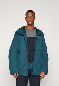 Veste imperméable bleu-vert avec une capuche, comportant plusieurs poches et une fermeture éclair. Portée par-dessus une chemise grise et des salopettes foncées.
