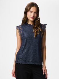 Barna hullámos hajú nő tengerészkék, ujjatlan csipkés felsőben, hullámos szegéllyel és fekete nadrágban, fehér háttér előtt áll.