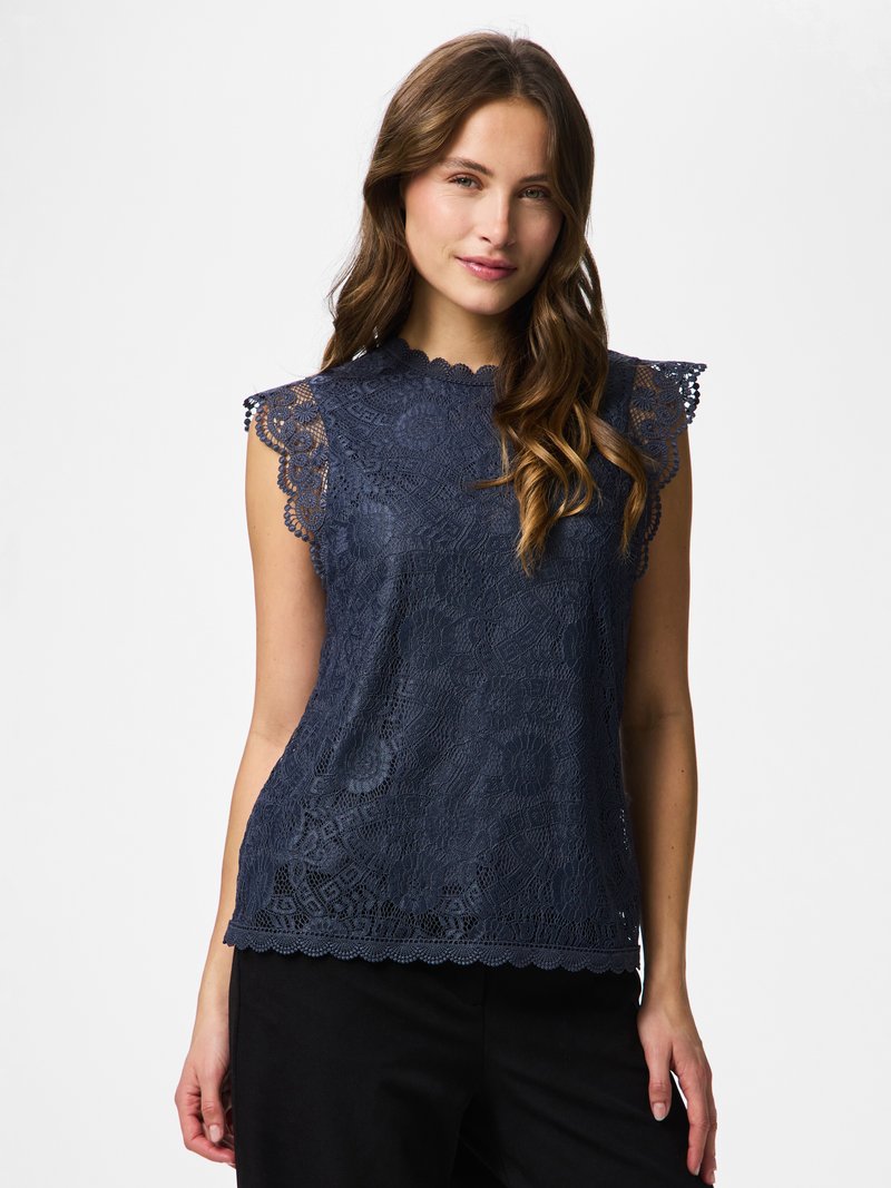 Barna hullámos hajú nő tengerészkék, ujjatlan csipkés felsőben, hullámos szegéllyel és fekete nadrágban, fehér háttér előtt áll.