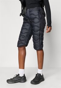 Calções pretos isolados com padrão acolchoado, apresentando um fecho éclair e bolsos laterais. Usados com sapatos pretos e meias brancas para contraste.